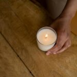 A close-up of a hand holding a lit candle on a wooden table, creating a cozy atmosphere.
