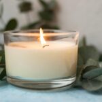 A lit candle in a clear holder surrounded by green leaves on a soft background, creating a calming atmosphere.