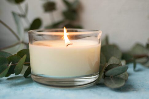 A lit candle in a clear holder surrounded by green leaves on a soft background, creating a calming atmosphere.
