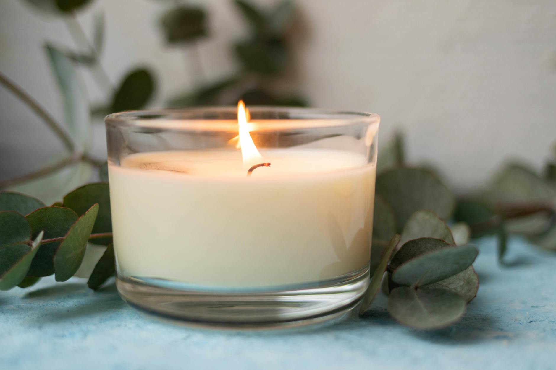 A lit candle in a clear holder surrounded by green leaves on a soft background, creating a calming atmosphere.