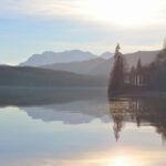 A tranquil mountain lake reflecting trees and mountains at sunset, creating a calm and peaceful scene.