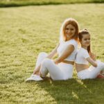A mother and daughter sit back to back in matching activewear on a sunny grass field, enjoying a summer day together.