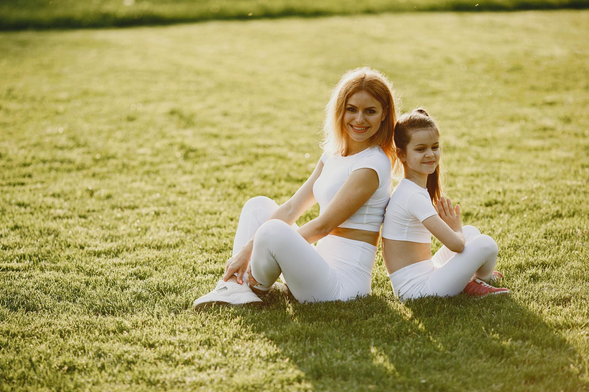 A mother and daughter sit back to back in matching activewear on a sunny grass field, enjoying a summer day together.