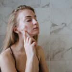Blonde woman using a jade roller for a relaxing skin care routine in a bathroom.