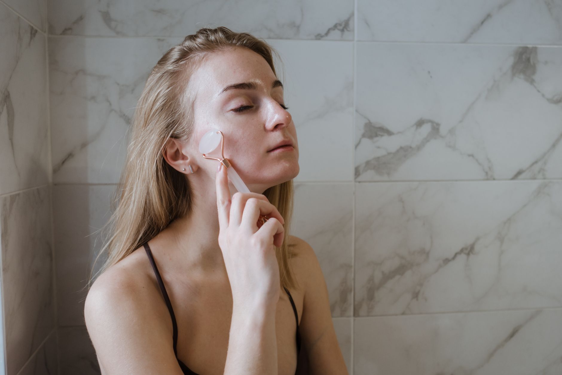 Blonde woman using a jade roller for a relaxing skin care routine in a bathroom.