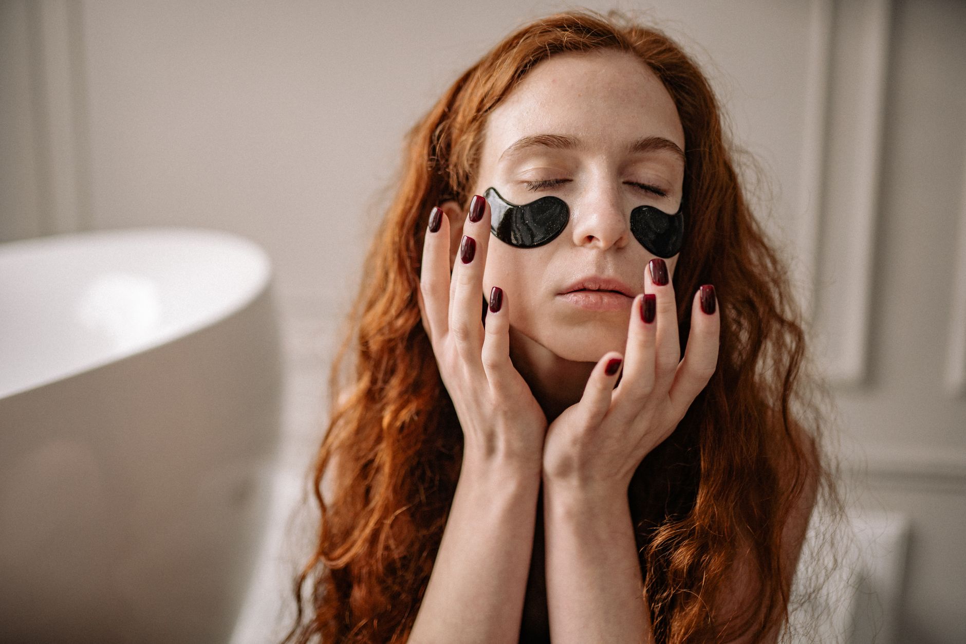 Woman with red hair enjoying a skincare routine with under-eye masks indoors.