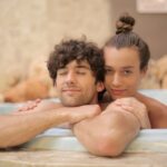 Young cheerful man and woman taking bath together enjoying holidays on blurred background of spa resort with marble interior and having romantic moments